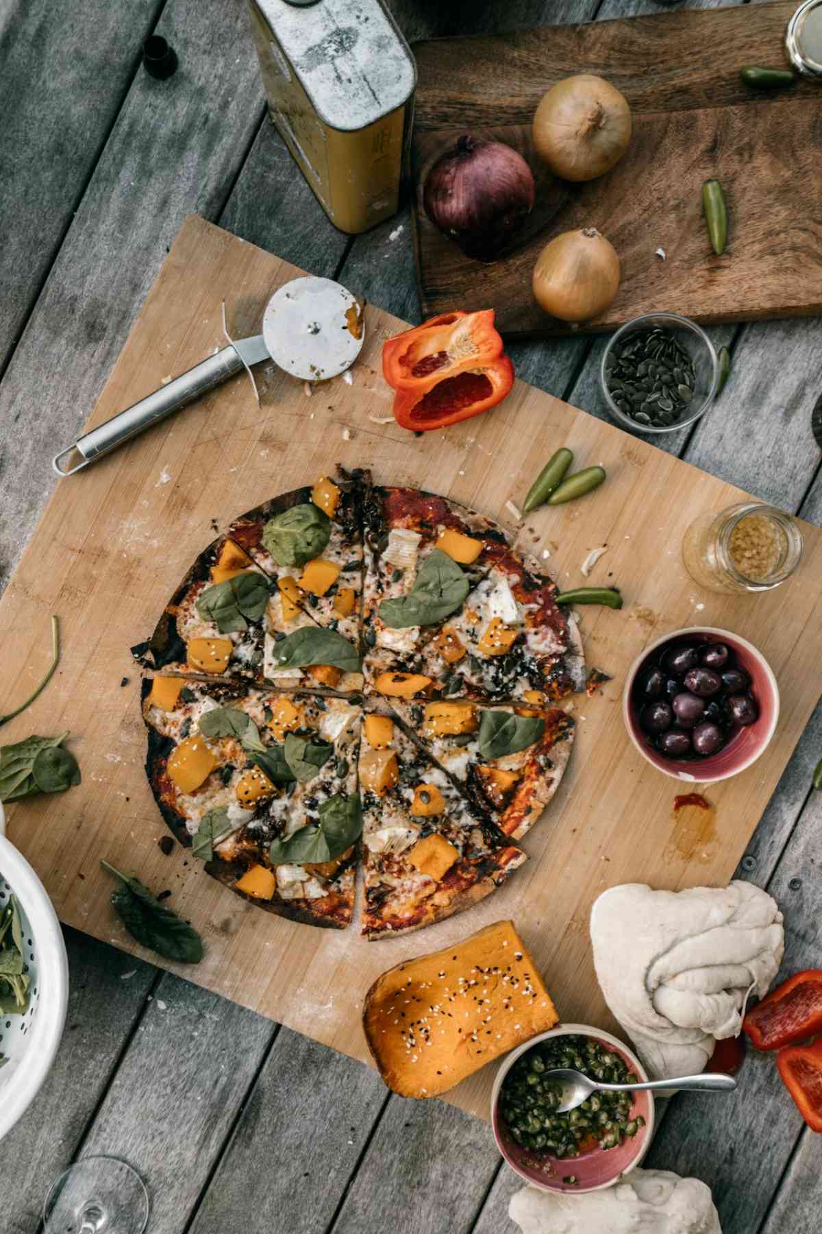 Overhead view of a sliced vegetable pizza on a wooden board, surrounded by pizza toppings, olives, a pizza cutter, and various vegetables on a rustic table in Temecula, CA. The perfect gluten-free and nightshade free pizza for women mindful of gut health.